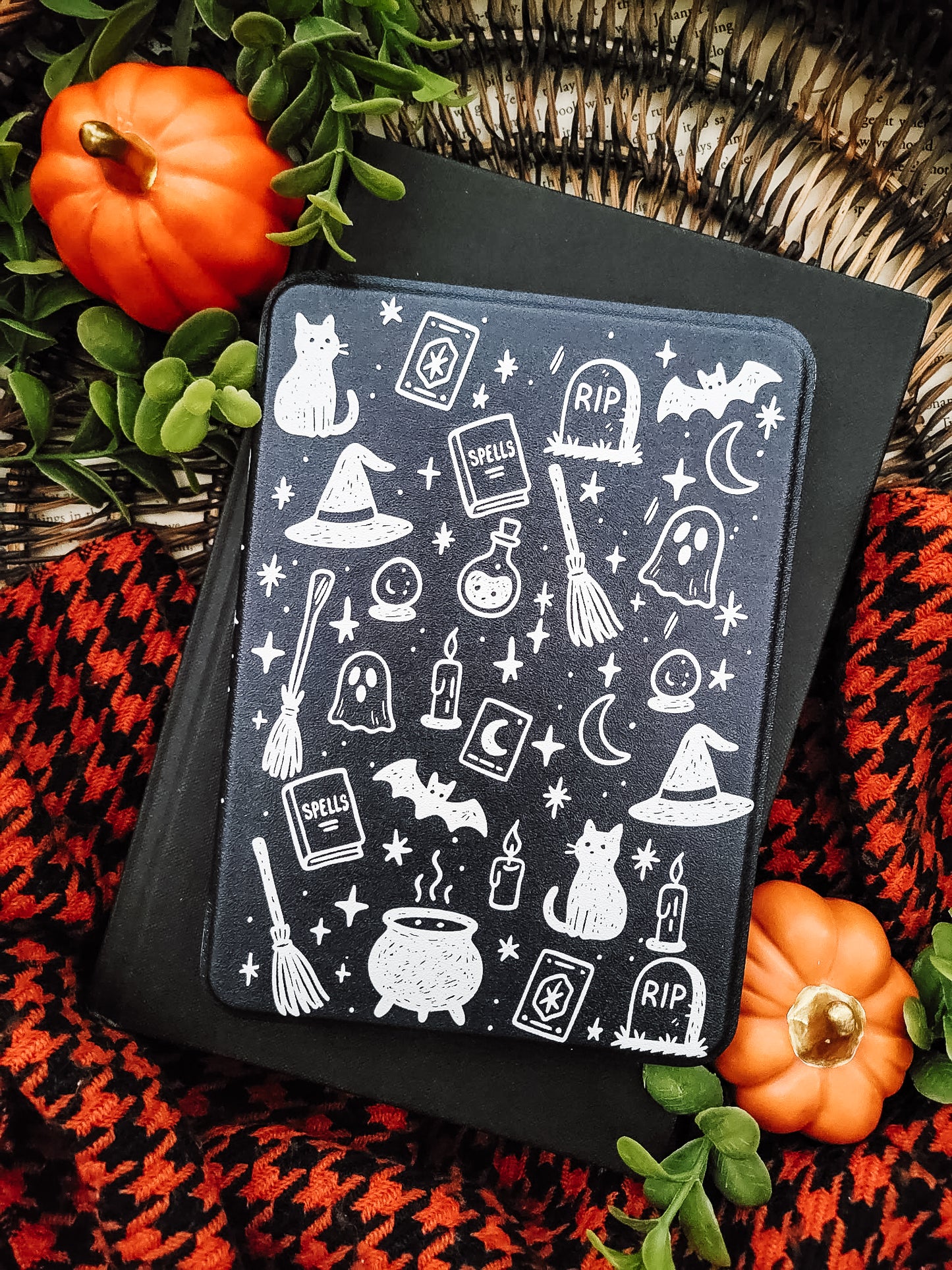 a Halloween-themed book cover with various spooky and supernatural elements, such as bats, skulls, pumpkins, and a cauldron, all depicted in white against a black background. The book cover is placed on top of a black book, surrounded by orange pumpkins and green leaves, creating a festive and eerie atmosphere.