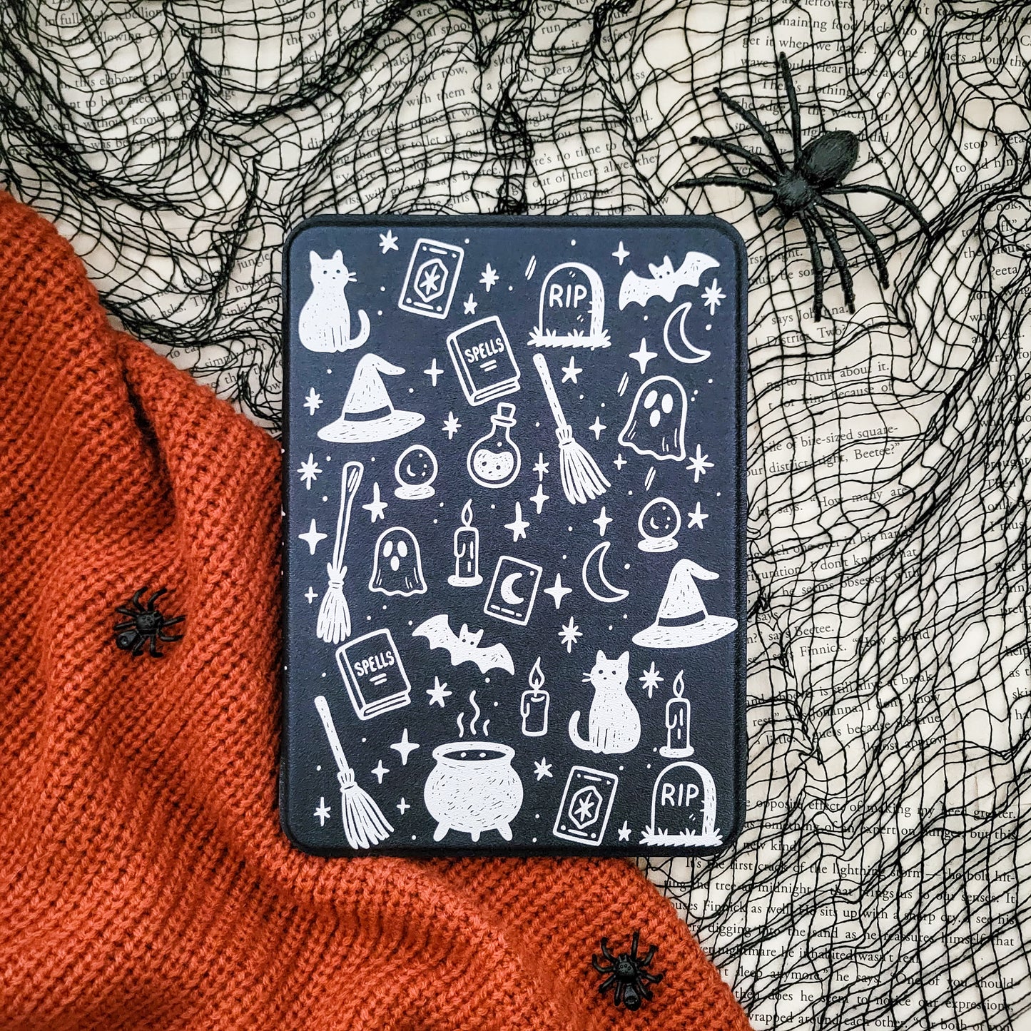 a Halloween-themed black case with white illustrations of various spooky and supernatural elements, such as bats, skulls, ghosts, and a spider, all set against a black background. The case is placed on a red knitted blanket, and there are black spider decorations on the blanket as well.