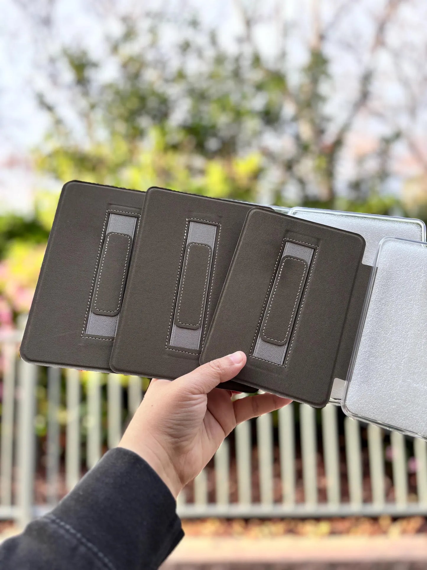 A hand holding a black tablet case with three compartments, against a blurred background of trees and foliage.
