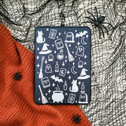 a Halloween-themed black case with white illustrations of various spooky and supernatural elements, such as bats, skulls, ghosts, and a spider, all set against a black background. The case is placed on a red knitted blanket, and there are black spider decorations on the blanket as well.