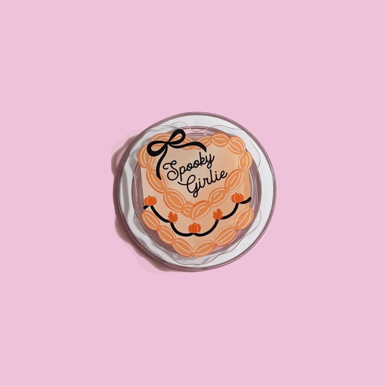 A cake with the text "Spooky Girlie" written on it, placed on a white plate against a pink background.