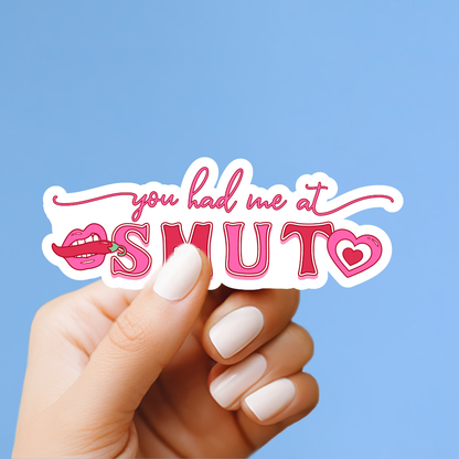 A hand holding a sticker with the text "you had me at smut" against a blue background.