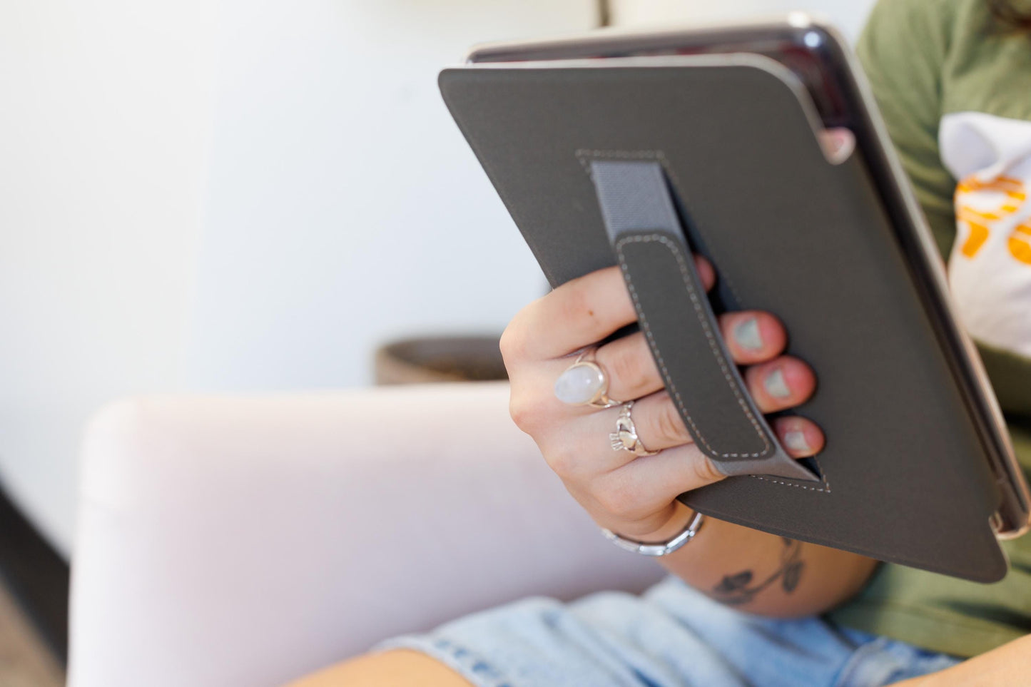 A person is sitting on a chair, holding a tablet device with a black cover and a gray strap.