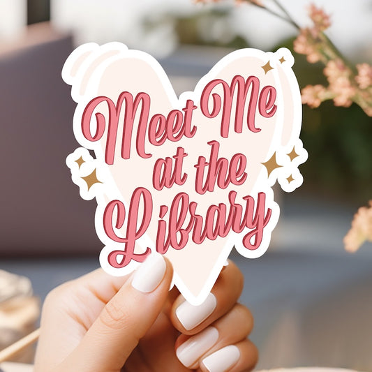 a person holding a heart shaped sticker with the words meet me at the library