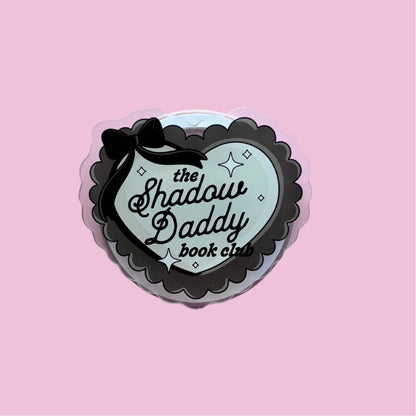 A heart-shaped sticker with the text "the Shadow Daddy book club" on it, set against a pink background.