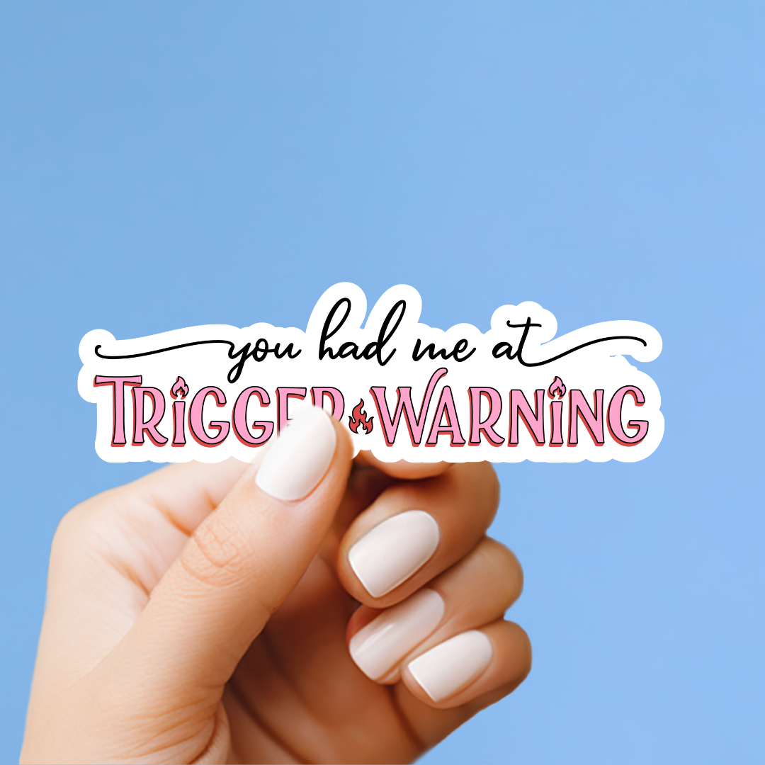 A hand holding a sticker with the text "you had me at trigger warning" against a blue background.